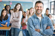 © Victor - Happy teacher standing in a class with crossed arms in front of his students and looking at the camera. Portrait of confident teacher with students studying in background.