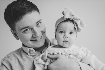 Wall Mural - Portrait of daddy with an baby girl with bow isolated in wall background. Cute baby in dads arms closeup. Dad holds in hands 6 month old little girl. Father hugging his daughter. Black and white photo