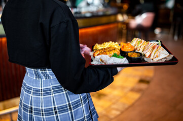 Wall Mural - Back view of a waiter holding a tray with a grilled sandwich, fries, and sauces in a cozy bar setting