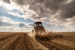 © oticki - Agricultural scene of a tractor plowing dry soil, kicking up dust with a dramatic sky overhead