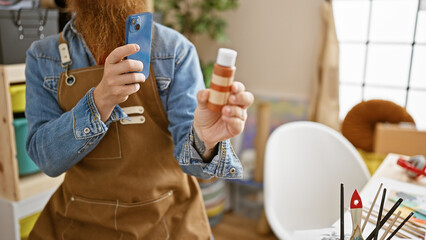 Canvas Print - Handsome, young, redhead man artist, clad in apron, taking smartphone picture to capture tube color for drawing in buzzing art studio.