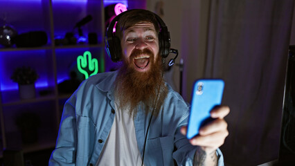 Wall Mural - Grinning gaming wizard! young irish redhead man streamer confidently hosts epic video call via smartphone in heart of night-dark gaming room