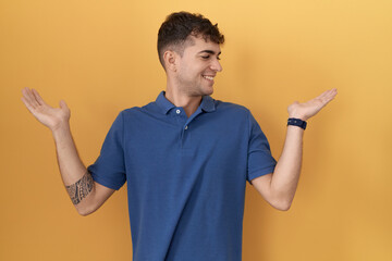 Canvas Print - Young hispanic man standing over yellow background smiling showing both hands open palms, presenting and advertising comparison and balance