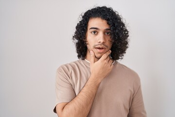 Sticker - Hispanic man with curly hair standing over white background looking fascinated with disbelief, surprise and amazed expression with hands on chin