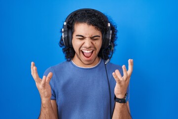 Sticker - Hispanic man with curly hair listening to music using headphones celebrating mad and crazy for success with arms raised and closed eyes screaming excited. winner concept