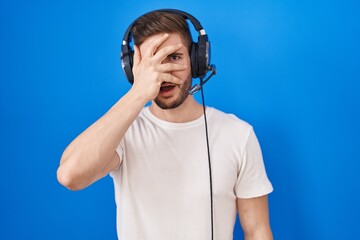 Poster - Hispanic man with beard listening to music wearing headphones peeking in shock covering face and eyes with hand, looking through fingers with embarrassed expression.