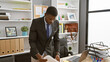 © Krakenimages.com - A professional african man reviews documents at a modern office setup with plants, shelf and leather bag in the background.