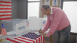 A mature man in a pink shirt writes at a polling station with us flags and a ballot box indicating a voting scene.