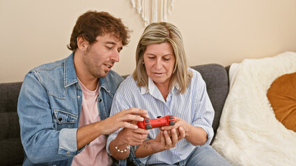 Wall Mural - Caucasian mother and son enjoying quality time together, casually sitting on living room sofa at home, teaching and learning to play a video game, emanating lovely expressions of teaching, gaming fun.