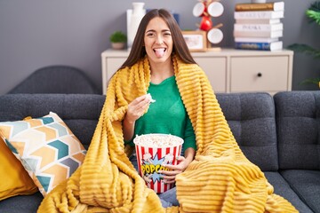 Sticker - Young hispanic woman eating snack sitting on the sofa at home sticking tongue out happy with funny expression.