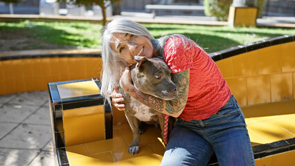 Canvas Print - Cheerful middle age woman joyfully hugging her grey-haired dog, while confidently sitting on a park bench, radiating pure happiness in the heart of a green outdoor city garden.