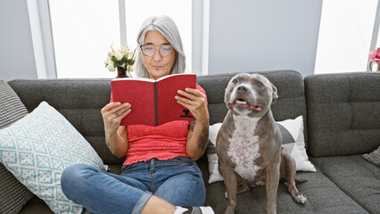 Poster - Smart grey-haired middle age woman comfortably reading literature with her dog on the sofa at home, enjoying her leisure time in a relaxed lifestyle.