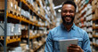A hardware warehouse is the setting for a laughing, smiling salesman who stands while reviewing supplies on his tablet.
