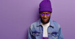 A man wearing a purple beanie and glasses is holding a cell phone. He is looking at the camera with a serious expression. man with phone on purple background