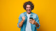 an exuberant young person with curly hair, holding a smartphone and seemingly shouting or cheering with a bright yellow background