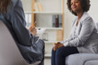 Female psychologist or therapist with clipboard and pencil in hands sitting on chair and talking to young African American woman patient on sofa. Therapy, psychotherapy concept