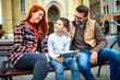 © Mediteraneo - Family sitting on bench, relaxing using gadget, browsing internet.
