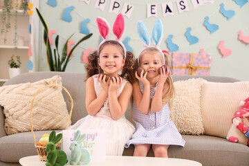 Wall Mural - Cute little girls in bunny ears with chocolate Easter eggs sitting on sofa at home