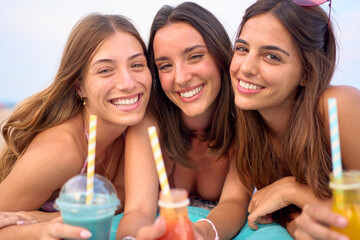 Wall Mural - portrait of three caucasian girls looking at camera and smiling. They drink natural smoothies. These attractive women look carefree and enjoy their vacation during the summer posing outdoors