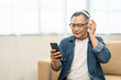 © Chanakon - Mature lifestyle relaxing on sofa in the morning. Happy Asian senior old man listening music with smartphone put on headphone by streaming music online. Choosing song by cell phone