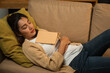 © Chanakon - Sleeping Asian beautiful woman lay down on cozey couch sofa while reading book in living room with warm light at night. People female reading book until fall asleep stay home part time relax at home
