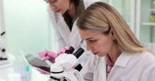 Woman looks in microscope while colleague inputs data in tablet