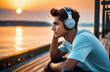 A young guy sits on a pier by the sea at sunset and listens to music with headphones
