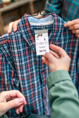 Wall Mural - Close up of unrecognizable female customer hand holding label of plaid shirt on clothing store. Woman employee showing tartan shirt on hanger to client in a shop. Selective focus on apparel label.