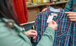 © David Pereiras - Close up of unrecognizable female customer looking label price of plaid shirt on second hand store. Woman employee showing discount clothes to client in a shop. Sustainable apparel shopping concept