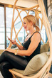 © carlesmiro - Confident satisfied young female entrepreneur in trendy informal outfit sitting in hanging wicker chair in creative workspace on mobile phone.