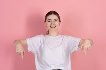 Wall Mural - Attractive Caucasian young brunette woman in a casual white t-shirt smiles, looks at camera and points down at empty advertisement copy space isolated on a pink studio background.