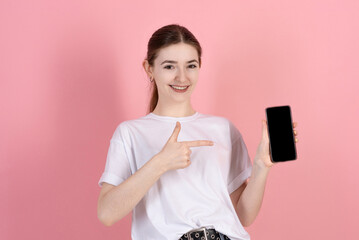 Wall Mural - Attractive Caucasian young brunette woman in casual white t-shirt points finger at blank mobile phone screen with copy space for advertisement isolated on pink studio background.