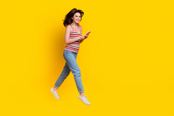 Canvas Print - Full body photo of satisfied cute girl wear striped tank fly hold smartphone walk to empty space isolated on yellow color background