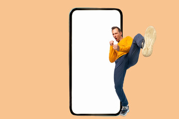 Full length profile shot of a young man doing karate jumping and gesturing while leaning on a big smartphone on isolated yellow background