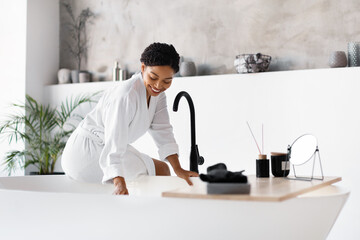 Wall Mural - Smiling black woman in bathrobe leaning over modern freestanding bathtub at home