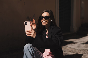 Wall Mural - Beautiful smiling fashionable businesswoman sitting on the stairs in front of old town and make selfie, hold mobile phone, look happy. Amazed woman make video call outdoors, hello hi gesture.