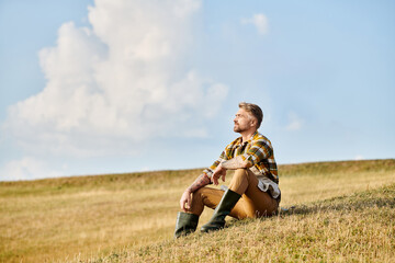 good looking dedicated man with beard relaxing on green spring field and looking away, modern farmer