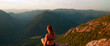 A young red-haired woman looks down from a mountain peak at several other mountains and forests. Meditation. Ecological concept.