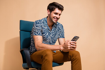 Canvas Print - Photo of happy young smiling businessman sits in conference room armchair browsing mobile phone isolated over beige color background