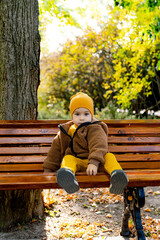 Sticker - A young child is sitting on a wooden bench in a park. The child is wearing a yellow hat and a brown jacket.