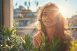 Close up of a smiling young woman standing at a beautiful modern terrace, she is spraying water on houseplants, lovely sunlit coming from her back...