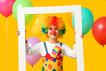 Poster - Funny little boy in clown costume with frame and balloons on yellow background. April Fools Day celebration