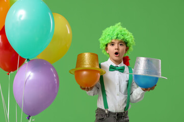 Poster - Surprised little boy in clown wig holding balloons with hats on green background. April Fools Day celebration