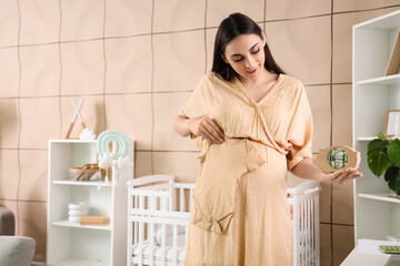 Wall Mural - Young pregnant woman with baby bodysuit and piggy bank at home. Maternity Benefit concept