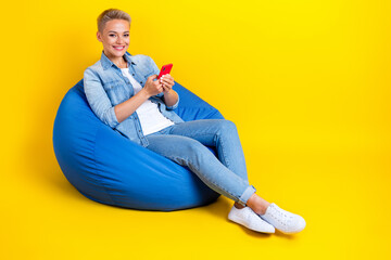 Wall Mural - Full size photo of pretty young girl sit bean bag hold device influencer dressed stylish denim outfit isolated on yellow color background