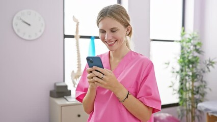 Poster - Blonde woman in pink scrubs smiling while checking phone in bright clinic room