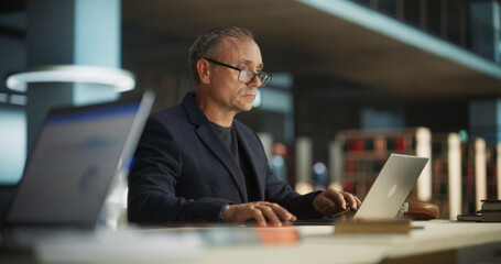 Knowledgeable Teacher Using Laptop Computer in a Library. Smart Middle Aged Male Educator Preparing Lectures for College Students, Checking Exams, Browsing and Researching Internet Articles