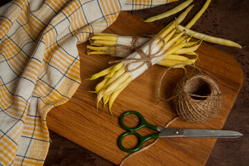 Cutting board with two bunch and several pods of flat runner bean pods heap with yellow kitchen towel, scissors and ball of thread on wooden background. .