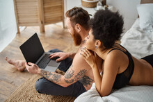 interracial couple lying on bed and enjoying cozy moment while watching movie on laptop together