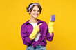 Young smiling happy woman she wear purple shirt rubber gloves do housework tidy up hold in hand point index finger on sponge isolated on plain yellow background studio portrait. Housekeeping concept.